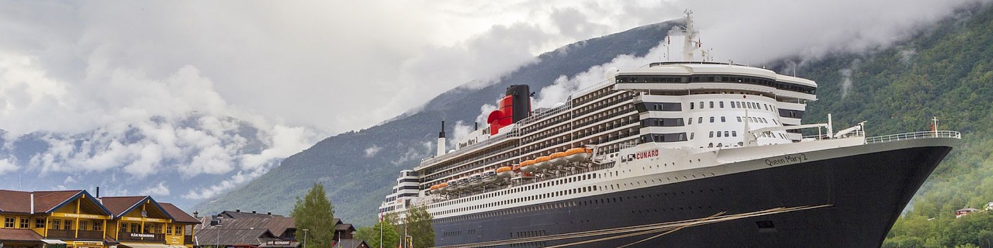 A large cruise ship docked by a scenic port, with mountains and clouds in the background, reflecting on calm water.