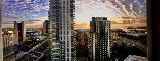 The image shows a cityscape at sunset, featuring tall buildings, a harbor, and a stunning sky with scattered clouds.