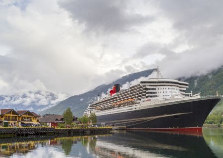 A large cruise ship docked near a quaint settlement with mountains and clouds in the background, reflecting in calm waters.