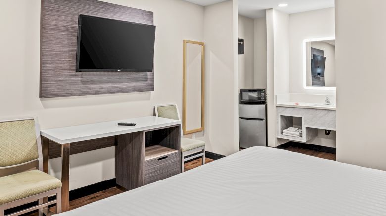 A hotel room with a bed, desk, chair, wall-mounted TV, fridge, microwave, mirror, and minimal decor, creating a cozy ambiance.