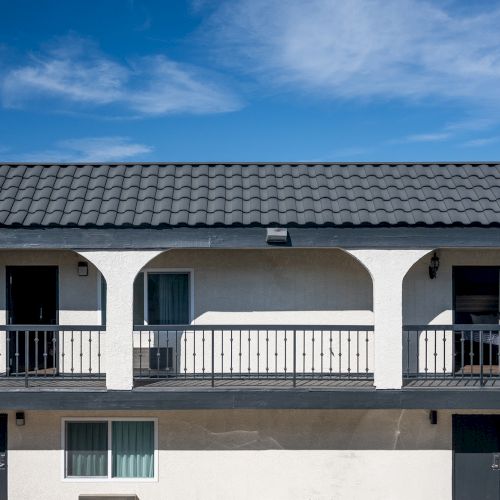 The image shows a two-story motel with a tiled roof, exterior corridors, and several doors and windows against a clear blue sky.