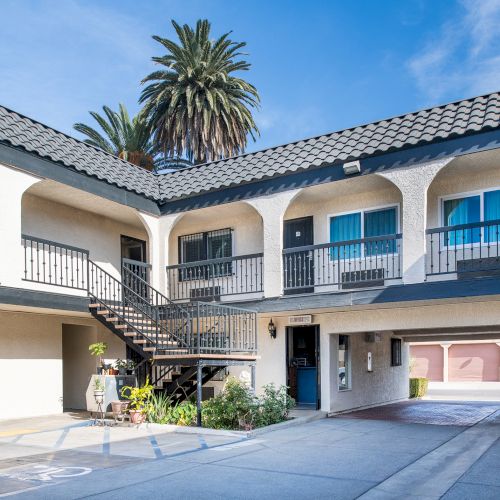 The image shows a two-story motel courtyard with stairs, palm trees, and a parking area under a clear blue sky.