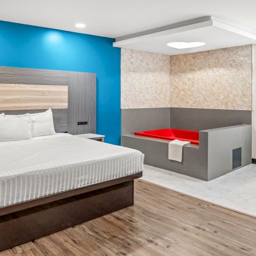 A modern hotel room with a bed, a red-tiled bathtub area, and a vanity mirror on the wall. The decor is sleek with wood and blue accents.