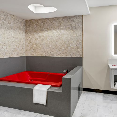 A modern bathroom with a red jacuzzi, a tiled wall, a white towel, a sink with a mirror, and a ceiling light shaped like a heart.