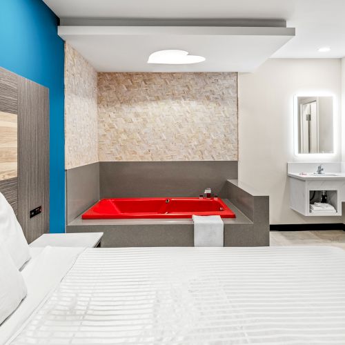 A modern hotel room with a bed, a red bathtub, a sink with a mirror, and a shower area is displayed.