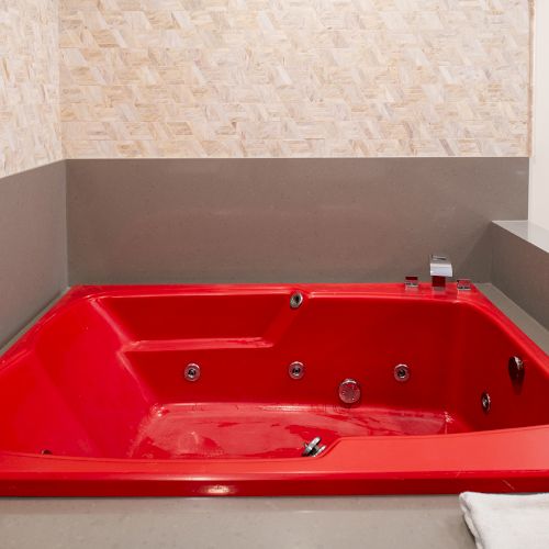 A red jacuzzi bathtub with jets is set in a tiled bathroom corner, featuring gray and beige walls, with a folded white towel nearby.