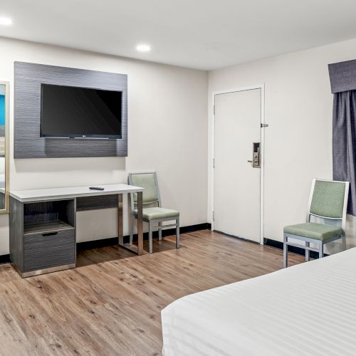 The image shows a modern hotel room with a bed, TV, table, chairs, a mini-fridge, and a microwave. The decor is minimalistic and clean.