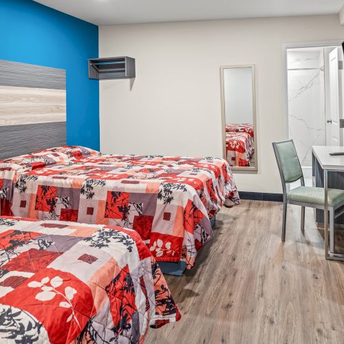 The image shows a hotel room with two beds, floral quilts, a desk, chair, TV, and mirror, featuring modern decor and wooden flooring.