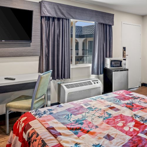 A hotel room with two beds, a flat-screen TV, desk, chair, mini-fridge, microwave, and window with curtains, featuring colorful bedding.