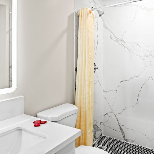 The image shows a modern bathroom with a sink, toilet, and a shower featuring a marble-patterned wall and a yellow curtain.