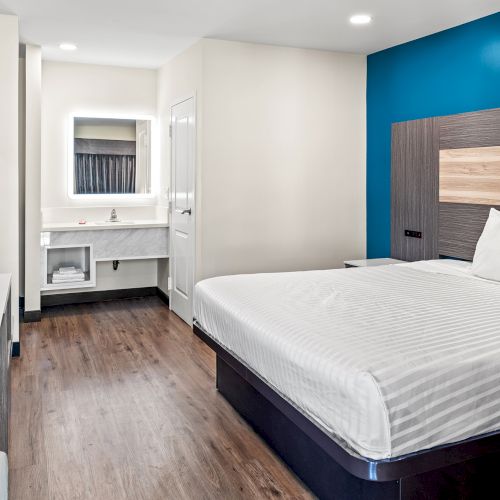 A modern hotel room features a bed, a wall-mounted TV, a desk, and a bathroom vanity, all highlighted by blue and white decor.