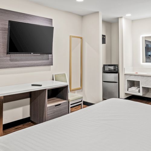 The image shows a hotel room with a bed, desk, chair, wall-mounted TV, mini fridge, microwave, and a mirror in a bright setting.