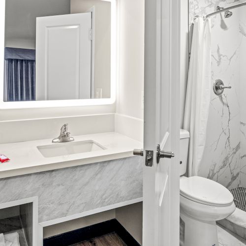 The image shows a bathroom with a sink, mirror, shower, and toilet; bright and modern design with marble accents and a small red item on the counter.