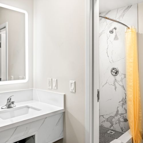 A modern bathroom with a lit mirror, sink, marble walls, yellow shower curtain, and toilet in a bright setting.