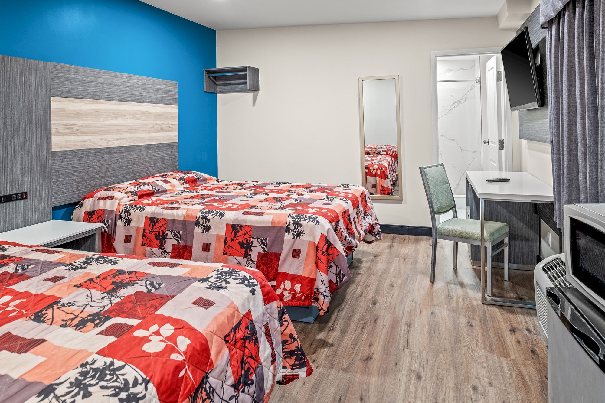 The image shows a modern hotel room with two beds, a desk with a chair, a wall-mounted TV, a full-length mirror, and vibrant bedding.