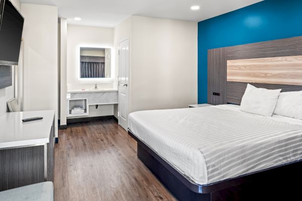 The image shows a modern hotel room with a large bed, blue accent wall, desk, wall-mounted TV, and a separate bathroom sink area.