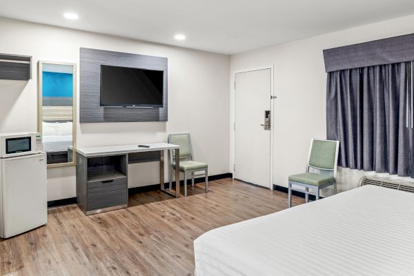 The image shows a hotel room with a bed, TV, desk, microwave, mini-fridge, and chairs, featuring wooden flooring and simple decor.