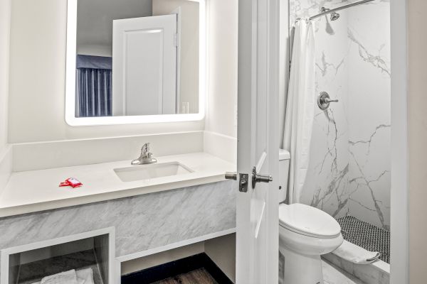 A bathroom with a sink, illuminated mirror, and marble shower. A toilet is next to the shower and a closed door is visible.