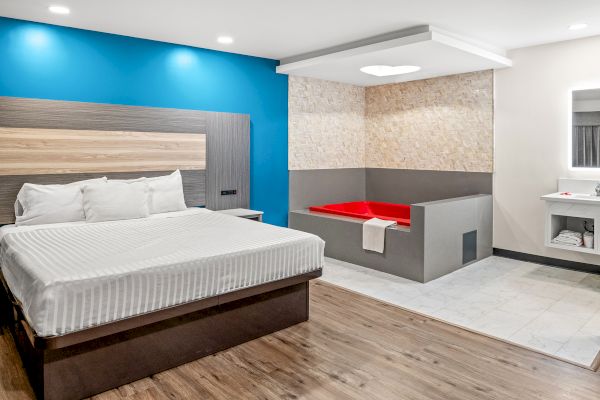 A modern hotel room with a bed, blue accent wall, wooden floor, and a corner jacuzzi surrounded by a tiled area.