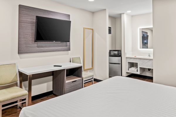 The image shows a hotel room with a TV, desk, chair, mirror, microwave, and mini-fridge. The room has neutral colors and modern decor.