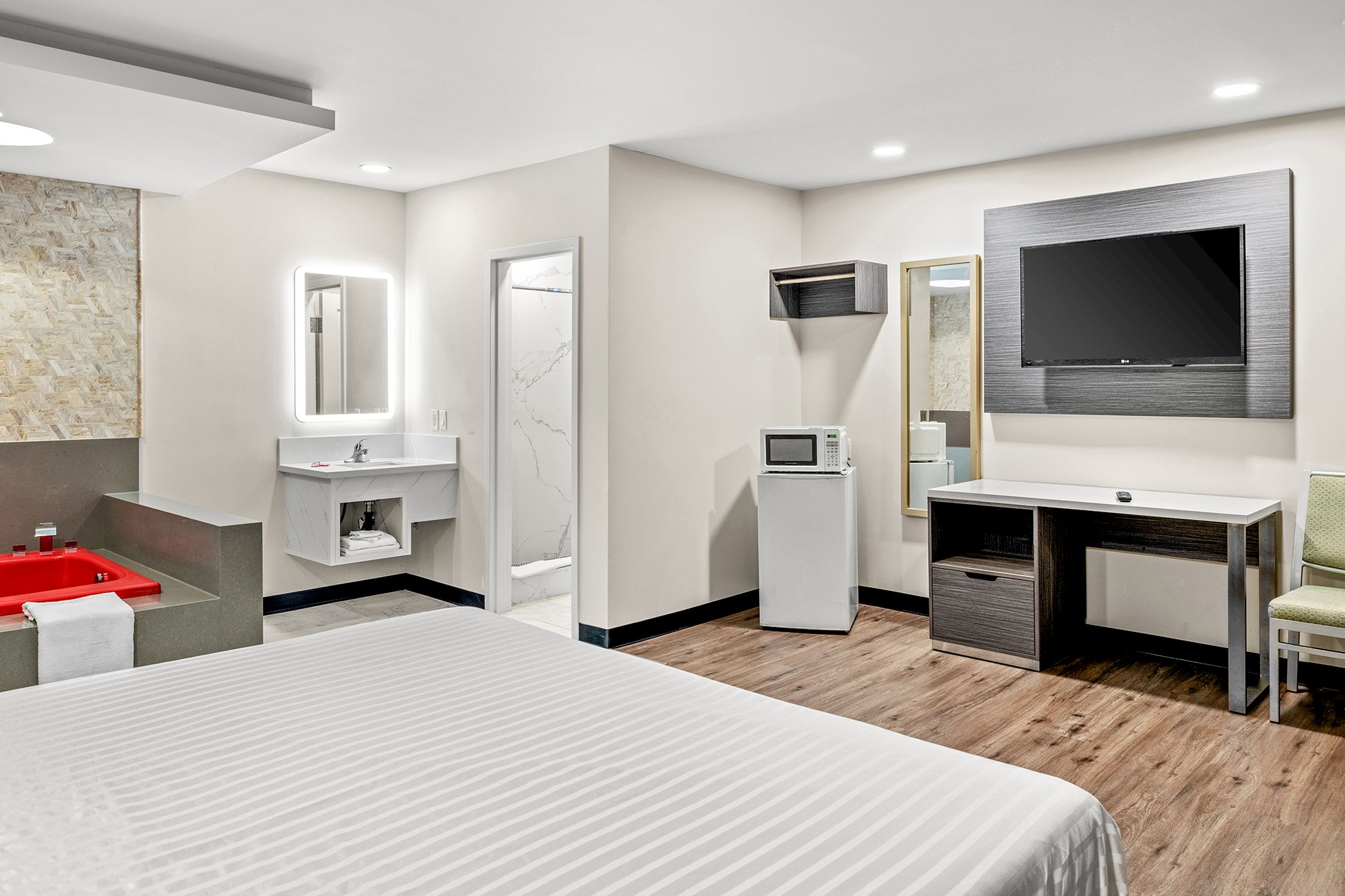 The image shows a modern hotel room with a bed, TV, desk, mini fridge, microwave, and a jacuzzi in the corner.