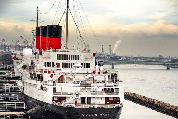 A large, historic ship docked at a harbor, surrounded by calm waters and a rock barrier. The ship has a red and black smokestack.