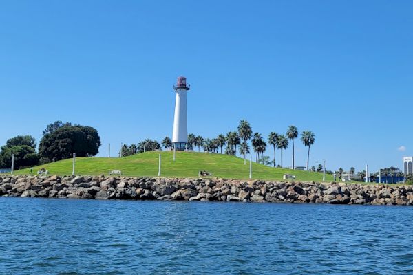 A lighthouse stands on a grassy hill with palm trees nearby, bordered by rocks and water under a clear blue sky.