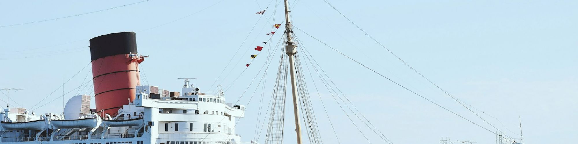 A large, historic ocean liner docked at a pier on a clear day, with a distinct red funnel and flags hanging from a mast, is shown.