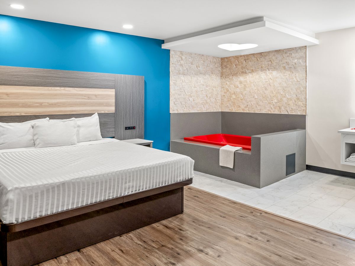 A modern bedroom with a large bed, vibrant blue accent wall, wooden flooring, and an elevated red jacuzzi next to the bed.