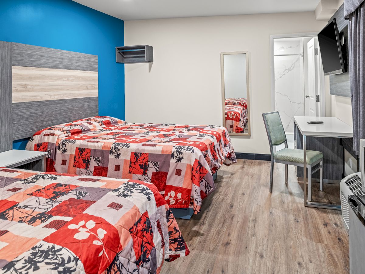 The image shows a hotel room with two beds, a desk, a chair, a TV, a microwave, and a bathroom door, featuring bright bedspreads and flooring.