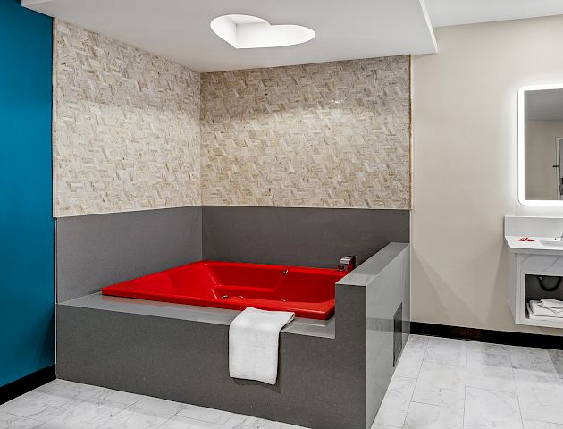 The image shows a room with a red bathtub, a towel, a wall mirror with lighting, and a sink on the side with modern decor.