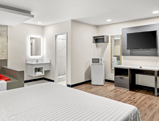 The image shows a modern hotel room with a bed, wall-mounted TV, desk, fridge, microwave, sink, and a red jacuzzi tub in the corner.