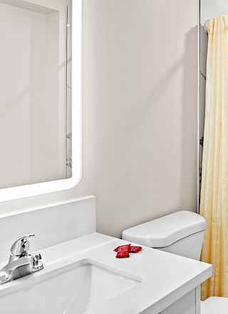 A modern bathroom with marble shower, white sink, wall mirror, and yellow shower curtain. A small red item is on the countertop.